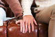 © artitwpd - Two people sit closely on a brown leather couch, one gently placing their hand over the other’s, capturing a tender moment of emotional support, comfort, and connection in a cozy indoor space.