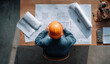© AndiAzis - Male construction worker wearing an orange safety helmet examining architectural blueprints at a wooden desk in a construction site
