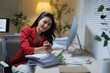 © Tj - Asian businesswoman working late at her desk, smiling as she sorts a large stack of reports and analyzes data on her computer with focus and professional dedication