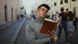 © Krakenimages.com - Young man reading a book standing on a busy city street with blurred people in the background in an outdoor setting, focusing intently on the pages