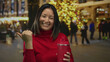 © Krakenimages.com - Woman in red sweater smiling outdoors with colorful city lights, holding a red bottle, and gesturing with her thumb on a festive urban street.