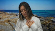 © Krakenimages.com - Woman standing on a beach with a pensive expression, wearing a white shirt, hands on heart, blue sea and rocky shore in background, summer day, evokes emotion, thoughtful at seaside