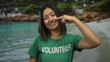 © Krakenimages.com - Young chinese woman smiling by the seaside, wearing a volunteer t-shirt, pointing to her nose, embodying the spirit of volunteering in a serene outdoor beach setting with clear blue water.