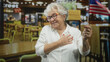 © Krakenimages.com - Woman holding small usa flag with right hand, left hand over heart, smiling in public building cafeteria; patriotic pride gentle remembrance.