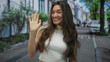 © Krakenimages.com - Young hispanic woman in white crop top waves hand and smiles on city street; warm greeting happiness.