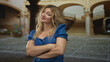 © Krakenimages.com - Woman blonde with arms crossed and bare forearms, smiling in building courtyard with arched colonnade; confidence resilience.