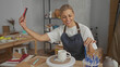 © Krakenimages.com - Woman holding smartphone with outstretched arm, gesturing to a ceramic mug on a potter's wheel in a pottery studio; creative focus.
