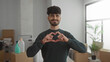 © Krakenimages.com - Young man making heart shape with hands in a new home surrounded by moving boxes and plants, expressing positivity and happiness in a bright living room setting.