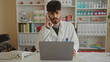 © Krakenimages.com - Young man in a clinic wearing a white coat talks on a phone while using a laptop in a professional workspace suggesting a busy work environment focusing on healthcare.