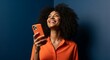 © nurman - Joyful young black woman with an afro hairstyle smiling while using her smartphone against a dark blue background.