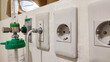 © Jamaludinyusup - close-up shot of essential hospital wall equipment, including an oxygen flowmeter with humidifier bottle, medical gas or data port, and European-style power sockets (Type F)