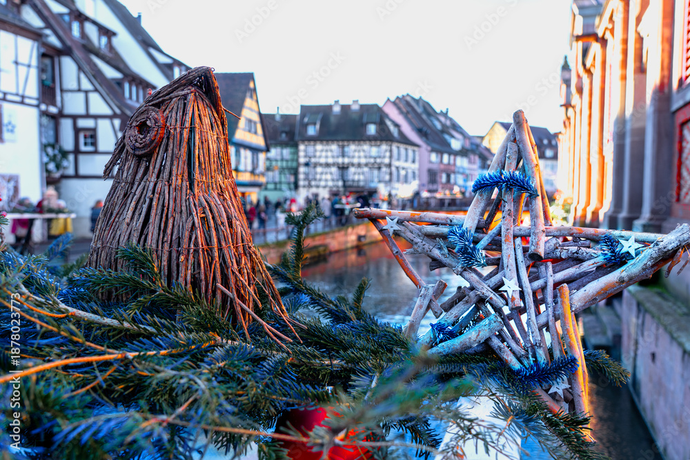 In the foreground, Christmas decoration with a star made of twigs and a bird's head made of rushes, with the Colmar canal very blurred in the background.