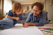 © Stockphotodirectors - A father and his young child are lying on a soft carpet, coloring and drawing with various pencils. The cozy living room has warm light and a comfortable sofa.