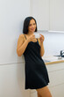 © makedonski2015 - A woman in a black dress stands in a white kitchen, smiling as she holds a white mug in her hands. She leans against a white cabinet
