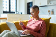 © Marko Geber - Older woman using laptop and credit card on sofa at home
