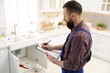 © New Africa - Plumber in uniform with clipboard writing in kitchen