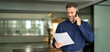 © insta_photos - Smiling middle aged senior professional businessman executive looking at documents talking on cell phone standing in office. Happy busy mature older business man holding papers making call at work.