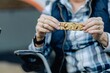 © Rawpixel.com - Person holding a granola bar, wearing a plaid shirt and vest. Close-up of hands with a focus on the snack. Outdoor setting with casual attire. Person eating proteinbar on camping trip and hike.