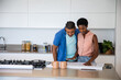© wavebreak3 - African American couple leaning over rolled-out blueprint on kitchen island, consulting smartphone