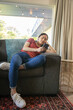 © wavebreak3 - Asian woman reclining on gray couch in living room leaning against teal pillow, using smartphone