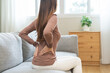 © KMPZZZ - Pain body muscles stiff problem, suffer asian young woman painful back, symptom injury ache from long study work, hand holding massaging or rubbing lower back hurt, sore sitting on sofa at home.