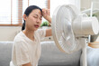 © KMPZZZ - Suffer summer heat wave stroke, hot weather, tired asian young woman sitting on couch in living room without air-conditioner, using electric fan, wave fan to cool for ventilation when high temperature