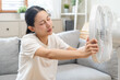 © KMPZZZ - Suffer summer heat wave stroke, hot weather, tired asian young woman sitting on couch in living room without air-conditioner, using electric fan, wave fan to cool for ventilation when high temperature
