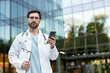 © Liubomir - Male doctor in a white lab coat and stethoscope holding a smartphone and clipboard, utilizing digital health technology for patient care and communication outside a clinic
