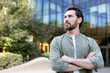 © Liubomir - Confident man with a beard and casual professional attire looking away, standing with arms crossed outdoors in a modern urban environment with a glass building and greenery