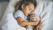 © Bonsales - Peaceful young child with curly hair sleeping soundly in a bright white bed, embracing a soft teddy bear