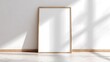 © oksanatukane - Wooden blank frame mock-up. Minimalist interior with a large empty wooden frame leaning against a white wall. Sunlight and soft diagonal shadows across the floor and the wall.