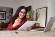 © fizkes - Happy excited young adult Indian girl in stylish glasses getting formal letter with good news, reading document at home work table with computer, making yes hand winner gesture