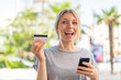 © luismolinero - Young blonde woman at outdoors buying with the mobile and holding a credit card with surprised expression