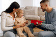 © Home-stock - Father giving red gift box to smiling little girl while woman looking at kid, family sitting on floor in living room, giving present to daughter
