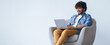 © Prostock-studio - A young man is sitting in a chair with a laptop on his lap. He is smiling while looking at the screen. This scene takes place in a bright and simple room during the day.