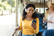 © Prostock-studio - Portrait of happy smiling black female passenger traveling in a train or bus, listening to music in wireless headset and using mobile phone, sending sms, standing in public transport near window