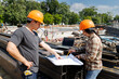 © skarie - Asian construction engineers discussing project blueprint on steel beams with laptop at site. Two civil architects checking building plans on metal stack at industrial construction work.