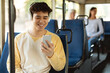 © Prostock-studio - Portrait of happy smiling Asian male passenger traveling in a train or bus, listening to music in wireless headset and using mobile phone, sending sms, sitting in public transport seat, enjoying ride