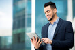 © Prostock-studio - Successful arab guy in formal wear standing by office building, holding digital tablet and smiling, looking for new business opportunities online, using business application, copy space, panorama