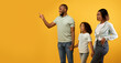 © Prostock-studio - Positive african american family of three looking aside, young man pointing finger at copy space, standing on yellow background, demonstrating empty place for your advertisement, panorama