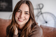 © luciano - Portrait of a smiling young woman with long brown hair sitting on sofa in a cozy modern living room. Natural light, warm tones. Concept of happiness, confidence, and everyday life