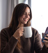 © luciano - Smiling girl sitting on couch holding mug of hot beverage, looking at her smartphone, enjoying aromatic herbal tea in morning routine, enjoying tech and social media
