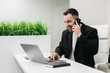 © wedmoments.stock - Male professional in a black blazer is engaged in a phone call while working on a laptop at a modern office desk with green plants in the background and a notebook nearby