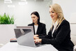 © wedmoments.stock - Two professional women in black suits working on laptops at a modern office desk with greenery in the background, engaged in a collaborative work environment