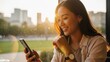 © Donnangelo - A young woman smiling and using her smartphone in a park with a city skyline in the background at sunset.