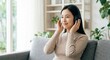 © Gayan - Peaceful Young Asian Woman with Headphones Relaxing on Sofa at Home, Smiling Adult Female Listening to Music in Modern Living Room, Wellness and Mental Health Concept, High-Key Interior
