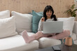 © wavebreak3 - Asian woman sitting on white sofa at home working on laptop with mug and smartphone