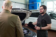 © pressmaster - Caucasian middle aged man in work overalls holding clipboard talking to bald Caucasian middle aged man, near open car hood in auto repair shop with vehicles in background