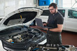 © pressmaster - Caucasian middle aged man wearing glasses standing in auto repair shop using laptop computer, while inspecting engine compartment of car with hood open in workshop environment