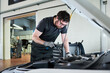 © pressmaster - Caucasian young adult man wearing glasses working on car engine in auto repair shop, using tool while inspecting under hood, focused on maintenance task in professional garage setting